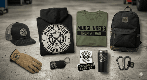Mudslingers Iron & Trail merch line laid out — black and olive tees, trucker hat, backpack, tumbler, gloves, stickers, patches, and key tag
